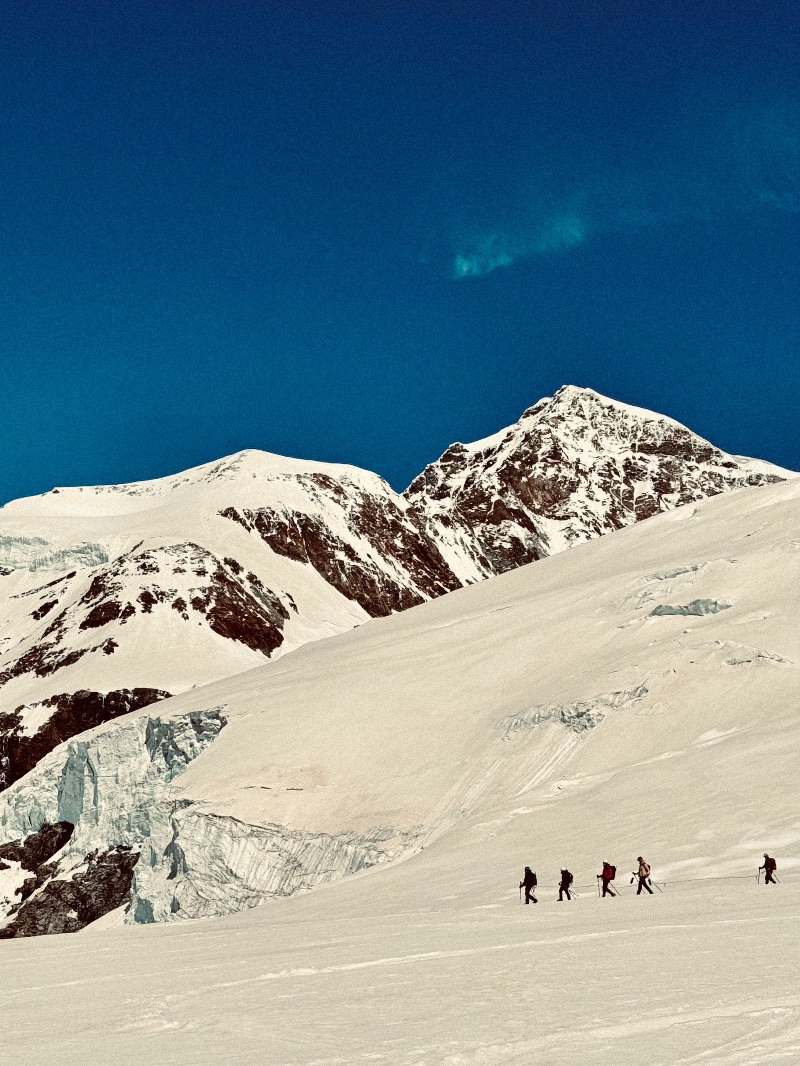 A group of people marching on snow with tall white mountains in the background.