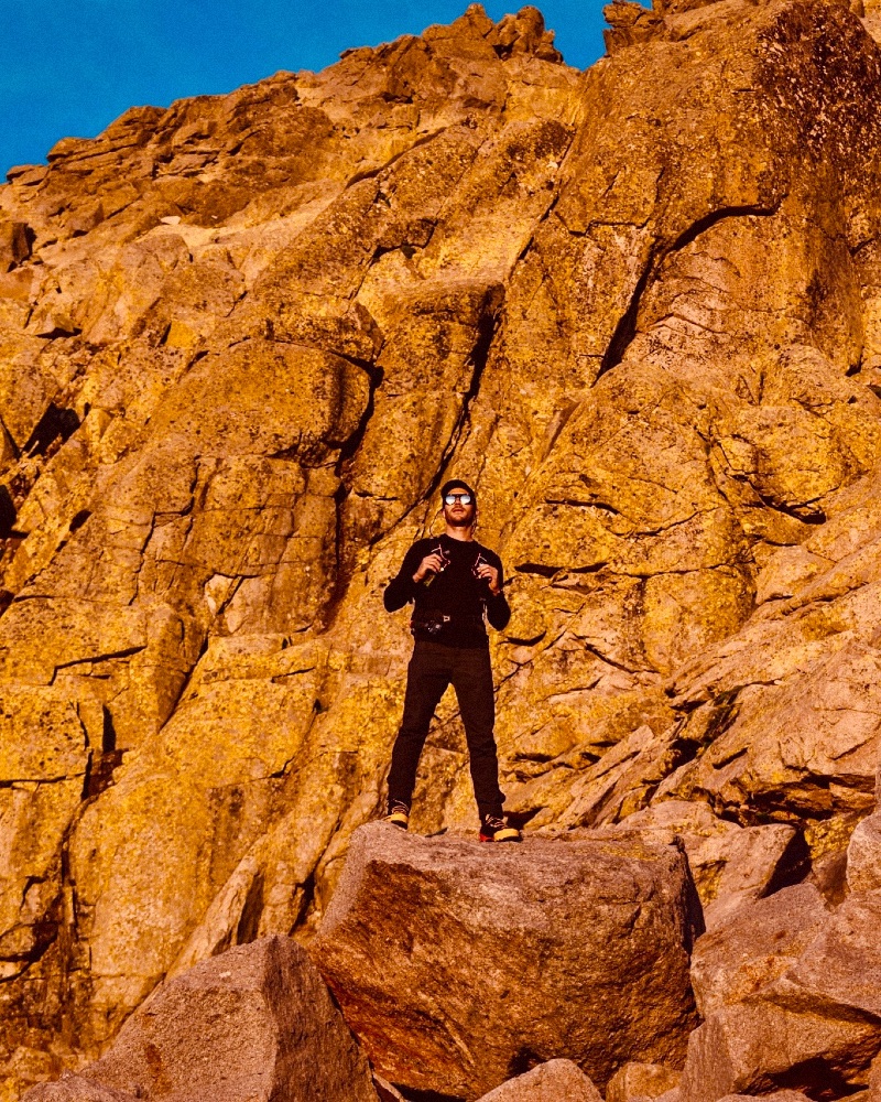 A man looking facing the sun set horizon with a dark orange rock wall behind him.