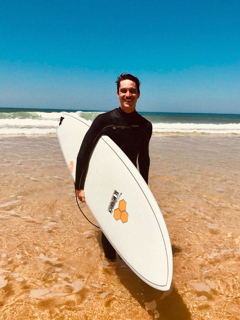 A man in a wetsuit holding a surfboard and laughing at the beach.