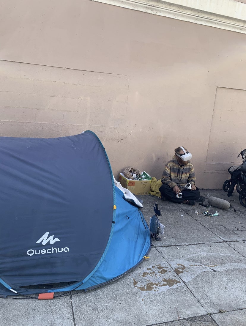 A homeless person sitting near their tent in the street with a pair of Oculus Quest 2 on their face.