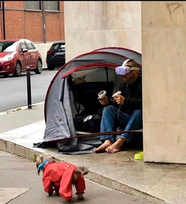 A homeless person sitting near their tent in the street with a pair of Oculus Quest 2 on their face.